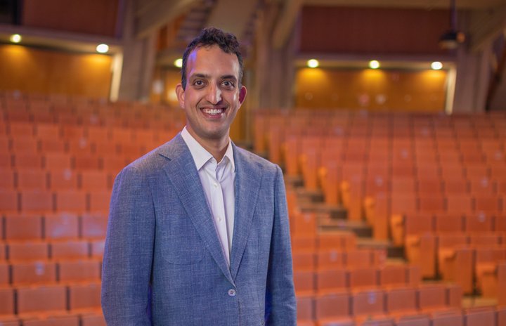 Prof. Dr. Siddharth Vedula is standing in a movie theatre wearing a blue suit and a white shirt.