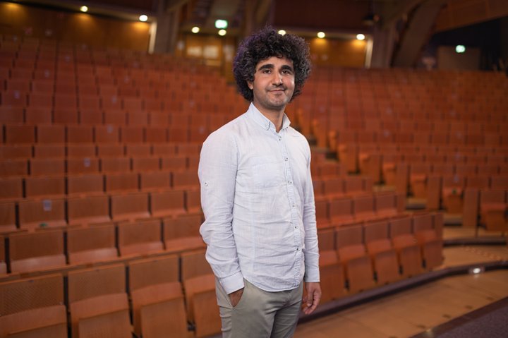 Phd candidate Cafer Bakac is standing in a movie theatre wearing a white shirt and beige trousers.