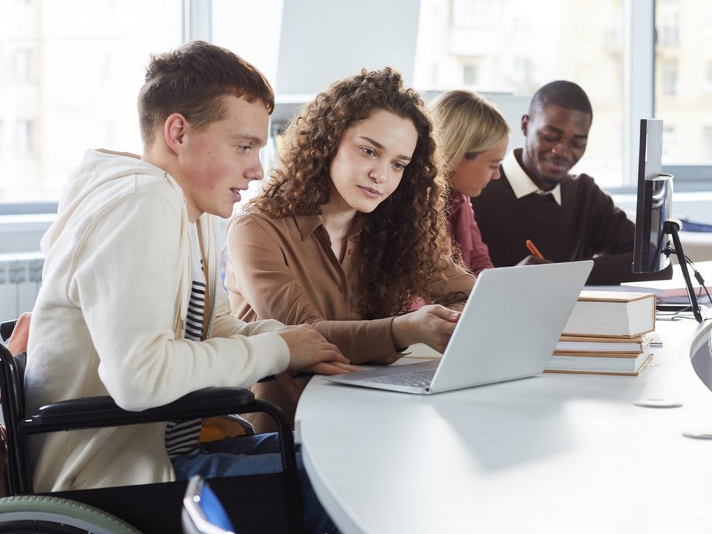 Side view at multi-ethnic group of students using laptop while studying in college, featuring boy using wheelchair