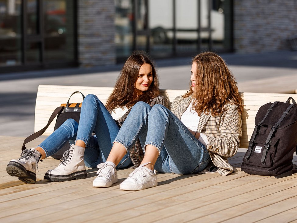 Two female students sitting on the ground, outside, chatting and smiling.