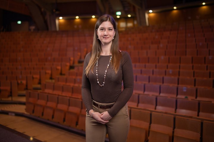 Researcher Dr. Sara Caprioli is standing in a movie theatre wearing a brown shirt with a necklace and beige pants .