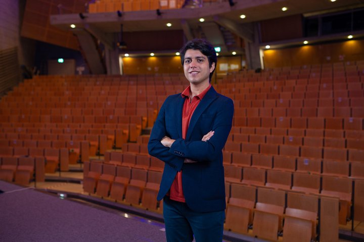 Phd candidate Baturhan Bayraktar is standing in a movie theatre wearing a suit and a red shirt. 