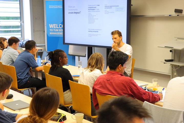 Lecture of the international summer school is sitting in fronmt of diverse international summer school students in a class room.