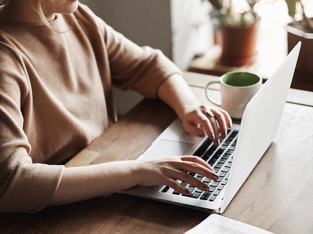 Vertical shot of determined and focused female student in glasses, sitting on wooden table, working with laptop and books, searching information 