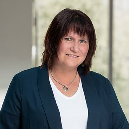 Portrait of our grade manager Ulrike Zerrahn. She has brown hair, wears a white shirt and a dark blue blazer and looks at the camera.