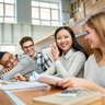 Multi-ethnic group of cheerful students sitting at desks in lecture hall and laughing, focus on two girls in front