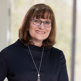 Portrait of our grade manager Annette Rank von Bronk. She has brown hair, wears a black pullover, a necklace and glasses and is smiling at the camera