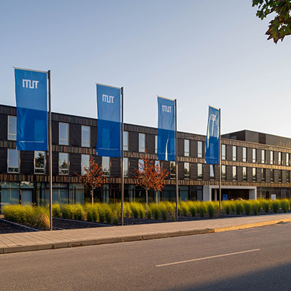 Modern brown flat building with three floors and four TUM flags in the foreground